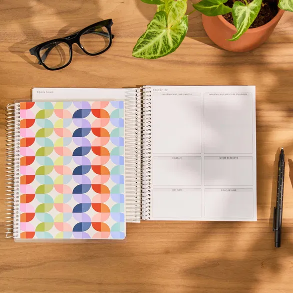 Open spiral-bound planner with colorful cover on a wooden desk with glasses and a plant.