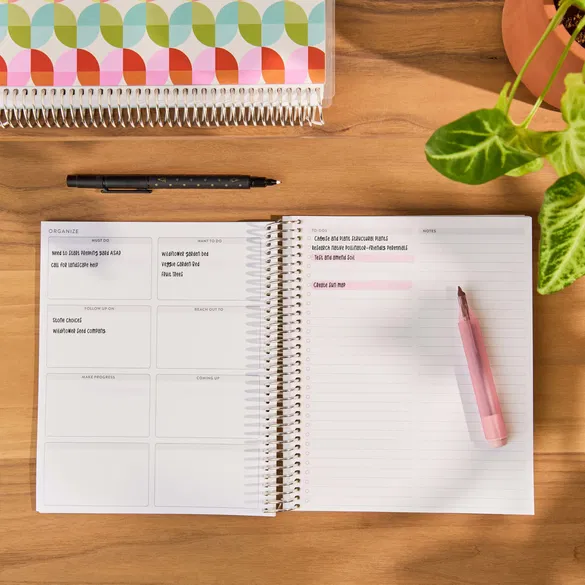Open planner with a pen on a wooden surface, next to a colorful notebook and plant.