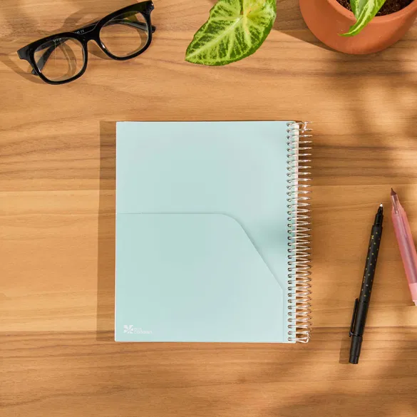 Spiral-bound notebook on a wooden surface with glasses, a plant, and pens.